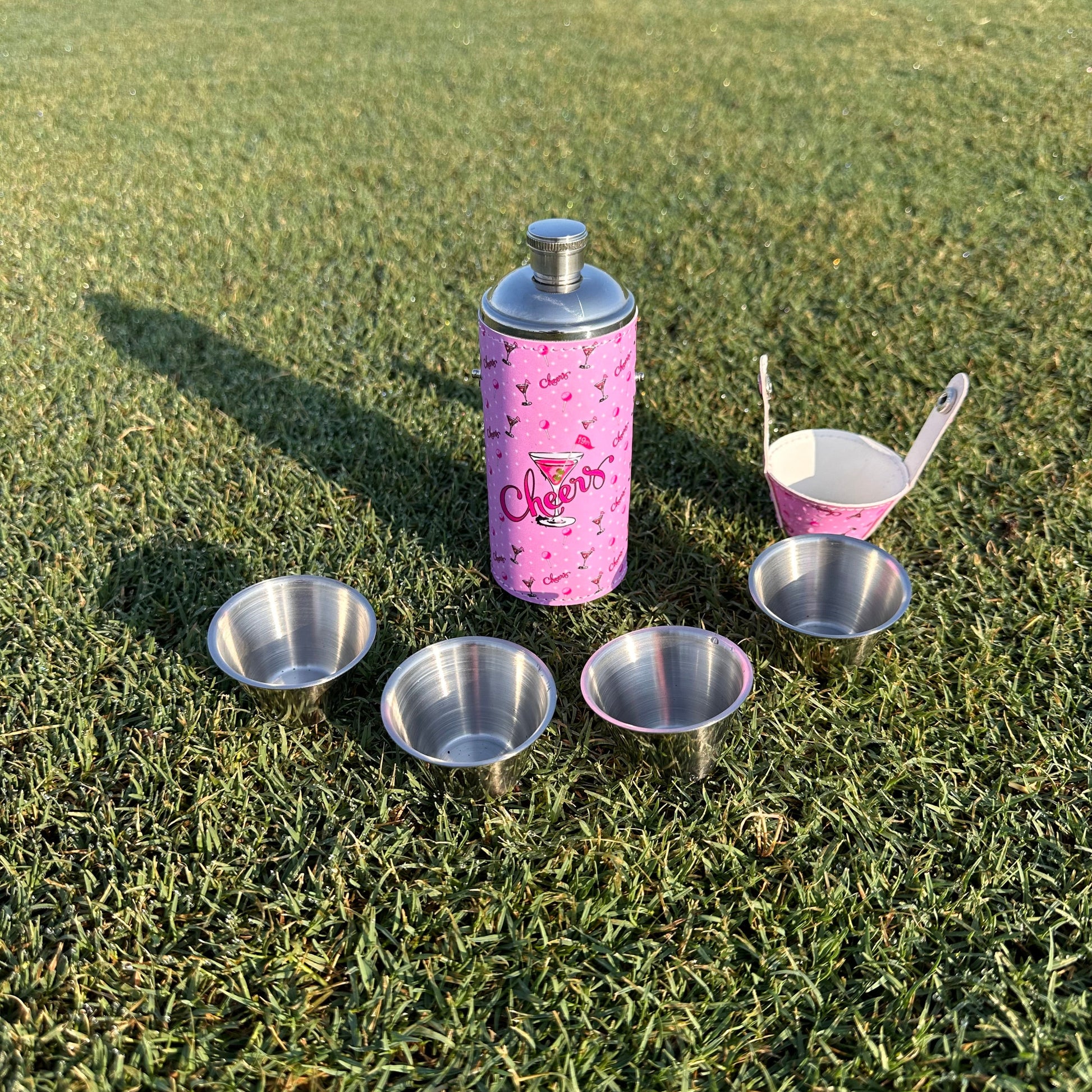 Pink flask bottle with silver shot glasses on green grass