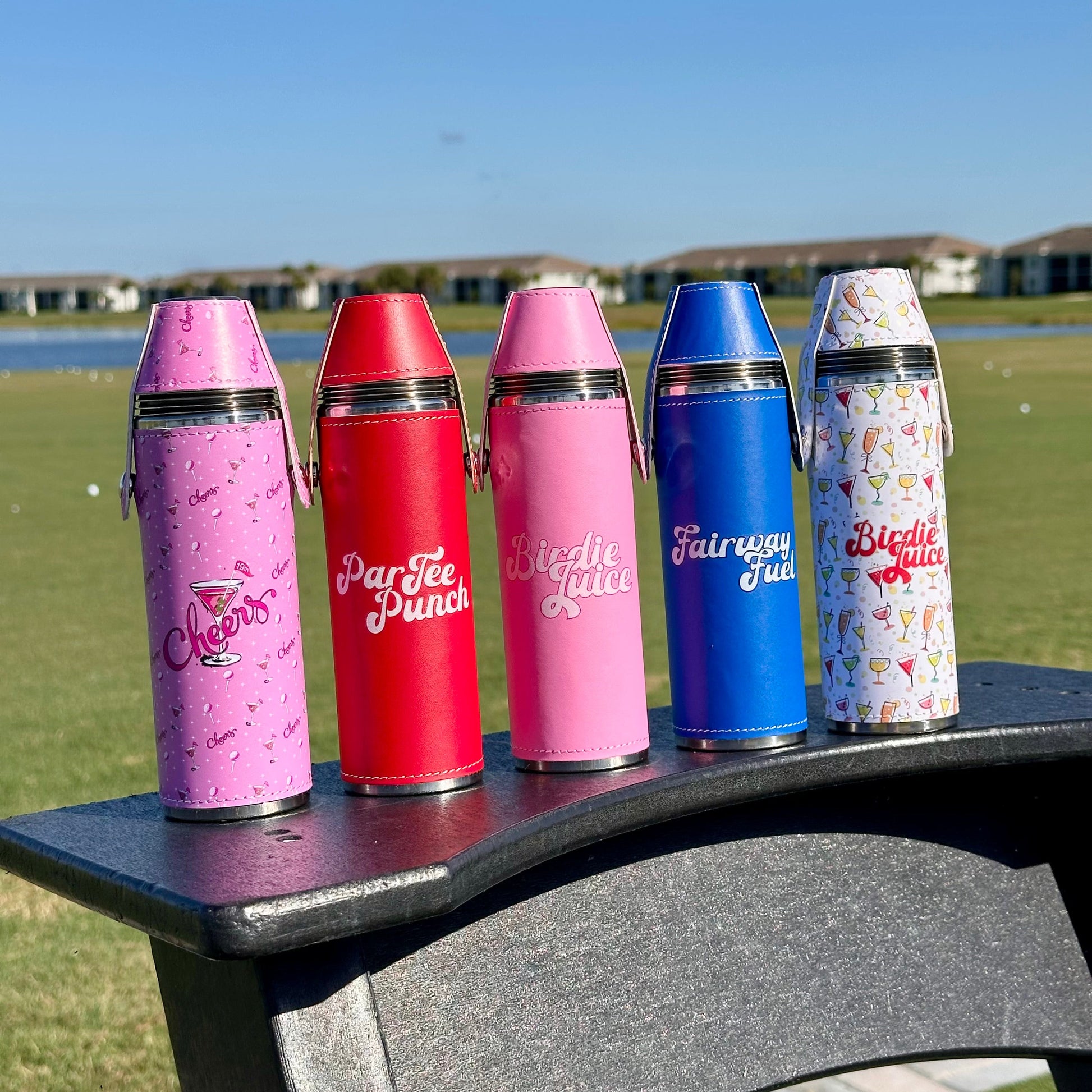 Five colorful flasks outdoors with a lake and buildings in the background.